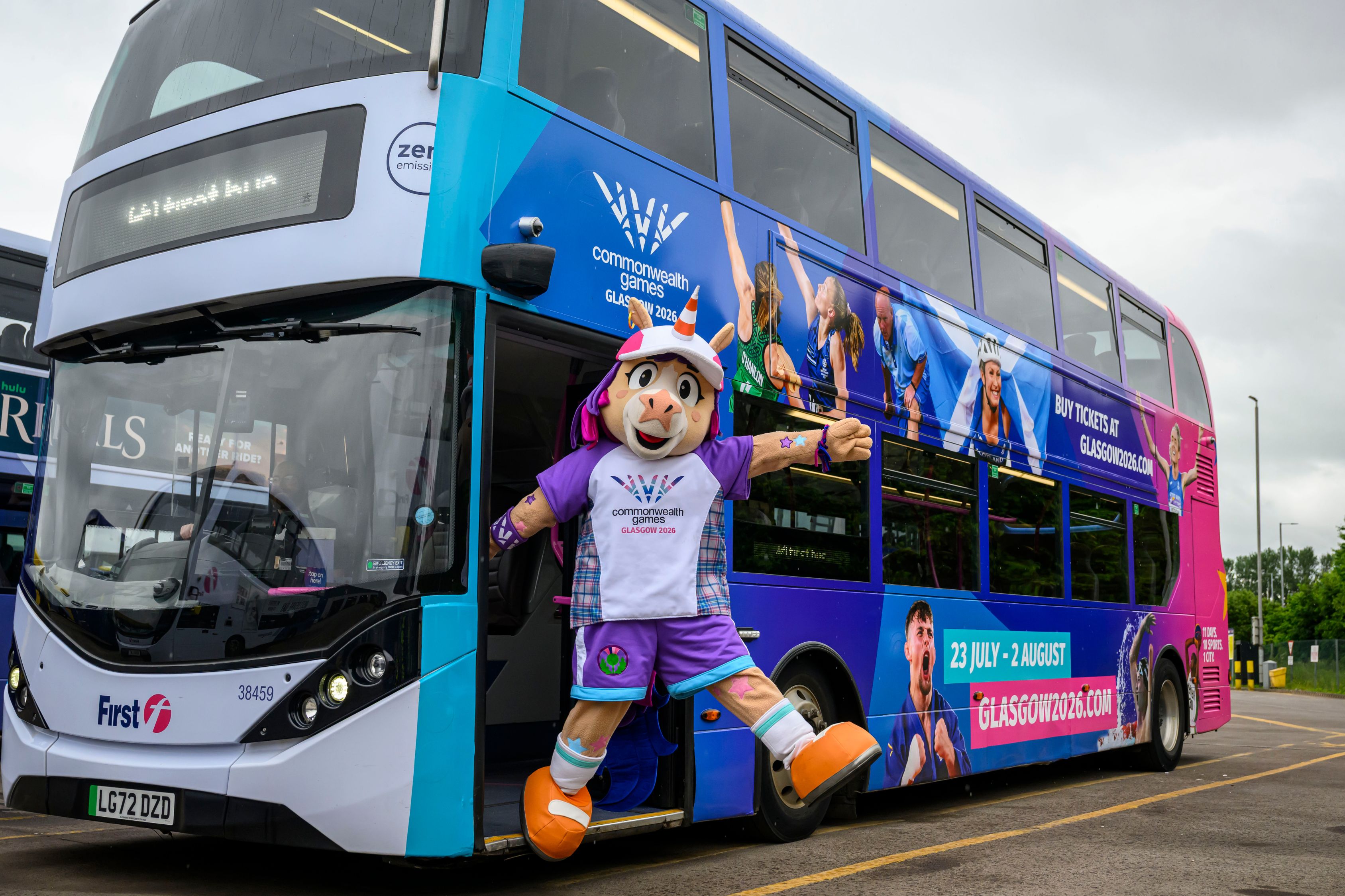 Finnie, the Glasgow 2026 Commonwealth Games mascot, steps out of a brightly branded double-decker First Bus featuring Commonwealth Games athletes and event artwork. The colourful bus, wrapped in blue, purple and pink Glasgow 2026 branding, is parked outdoors under a cloudy sky, promoting ticket sales for the Games taking place from 23 July to 2 August 2026.